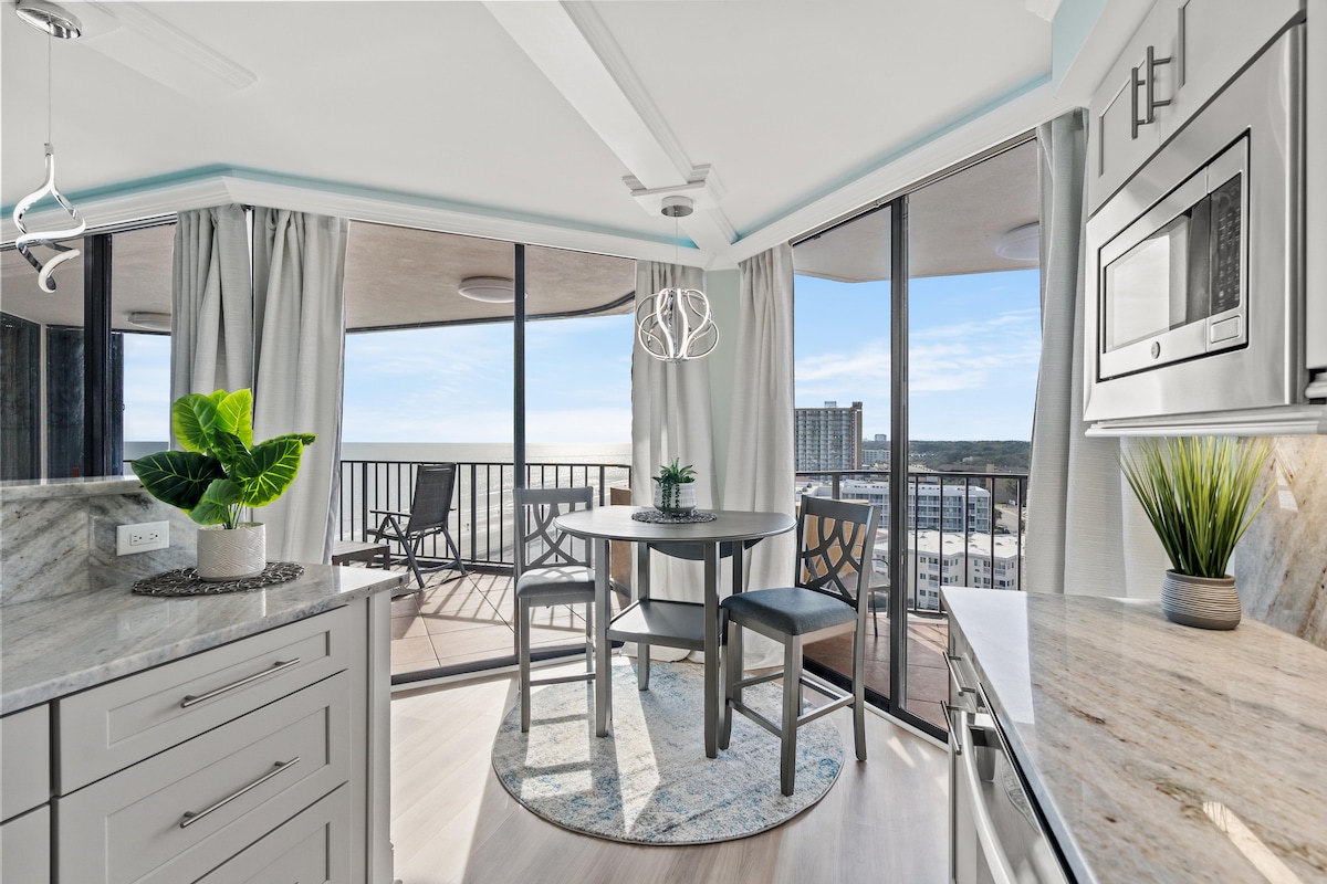 A modern dining area is positioned near large floor-to-ceiling windows, offering an ocean view. A round table is accompanied by high-backed chairs, surrounded by light-colored curtains that enhance natural light. Decorative plants are placed on the table and countertop, adding a touch of greenery.