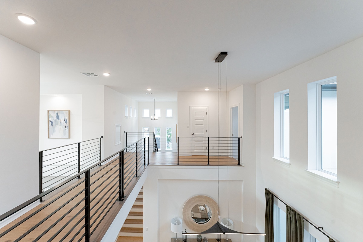 An airy interior space is presented with a two-story entryway, highlighting a modern railing along a spacious walkway. Natural light fills the home through numerous windows, while a circular mirror serves as a focal point in the welcoming area below.