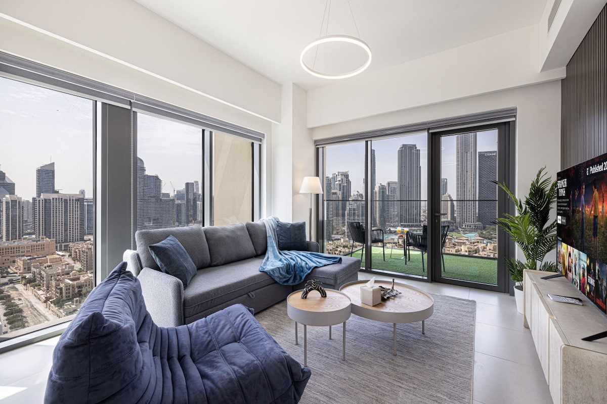 A modern living area showcases a spacious grey sofa adorned with a soft blue throw. Natural light fills the room through large windows, offering stunning views of the city skyline, including the Burj Khalifa. A round coffee table sits at the center, complemented by a cozy chair.