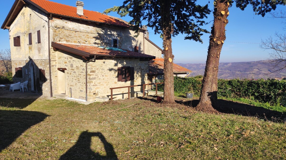 The exterior of a rustic stone house is shown, featuring a red-tiled roof and wooden shutters. The expansive lawn is bordered by trees, with a wooden walkway leading to the entrance. The surrounding landscape reveals rolling hills under a clear sky.