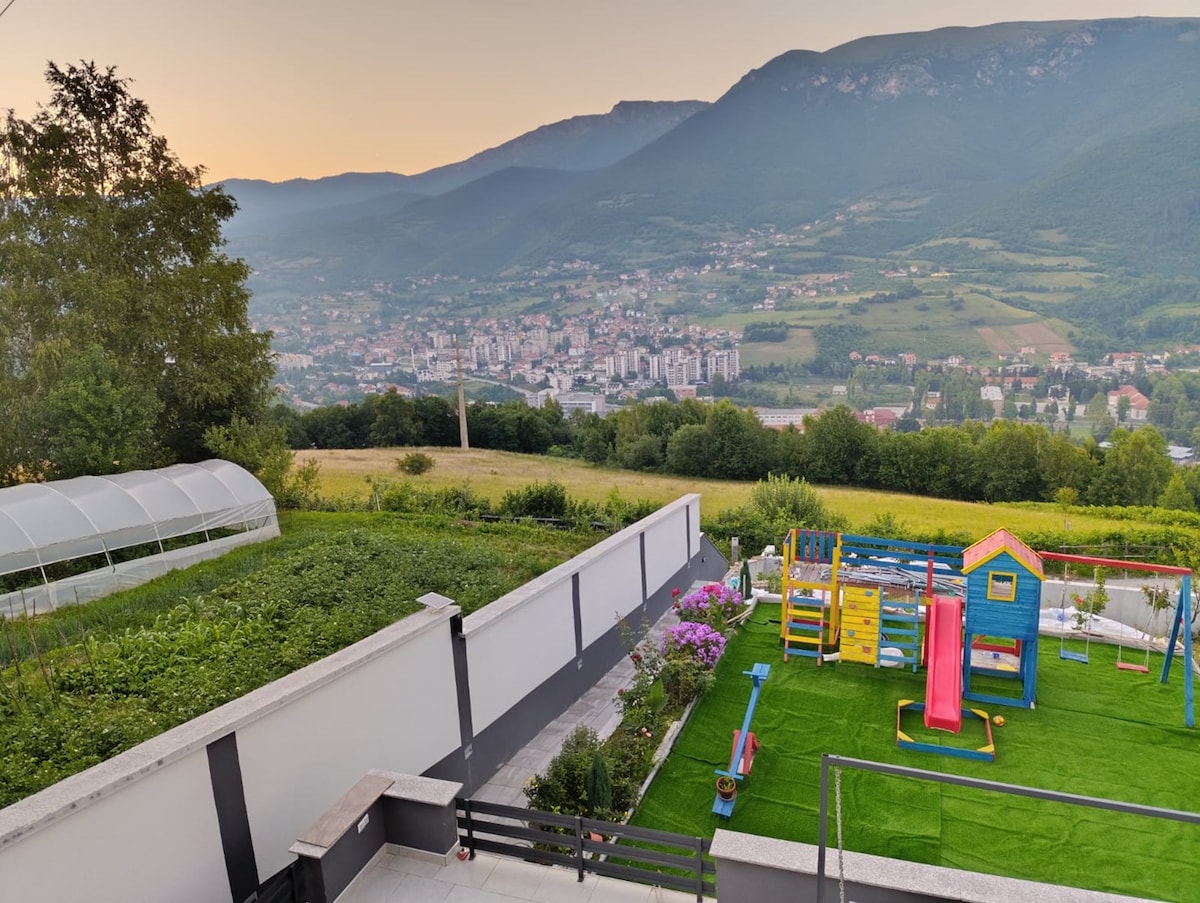 A vibrant children's playground is visible, featuring a slide, swings, and a playhouse. Lush greenery frames the area, while the stunning backdrop showcases mountains and the city of Travnik, highlighting the scenic beauty of the location.