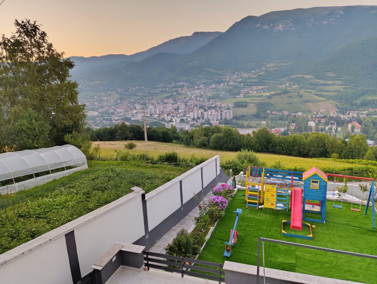 The image displays a vibrant children's playground with colorful equipment, including a slide and swings, situated on a grassy area. In the background, the scenic view of Travnik city and the surrounding mountains creates a serene landscape, framed by lush greenery.