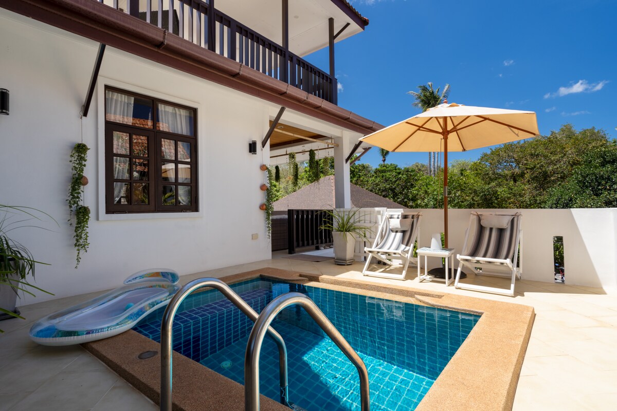 A private plunge pool is framed by a spacious patio area featuring two lounge chairs and a large umbrella. Greenery is visible in the background, while sunlight reflects off the clear water, enhancing the serene outdoor setting.