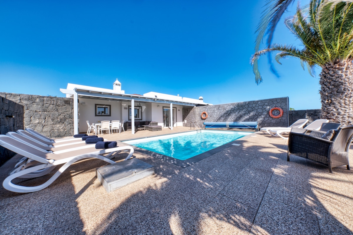 An outdoor space features a heated pool surrounded by sun loungers and chill-out sofas. The walls provide privacy, and an inviting seating area is positioned under clear blue skies. A barbecue area is visible, enhancing the appeal of this relaxing environment.