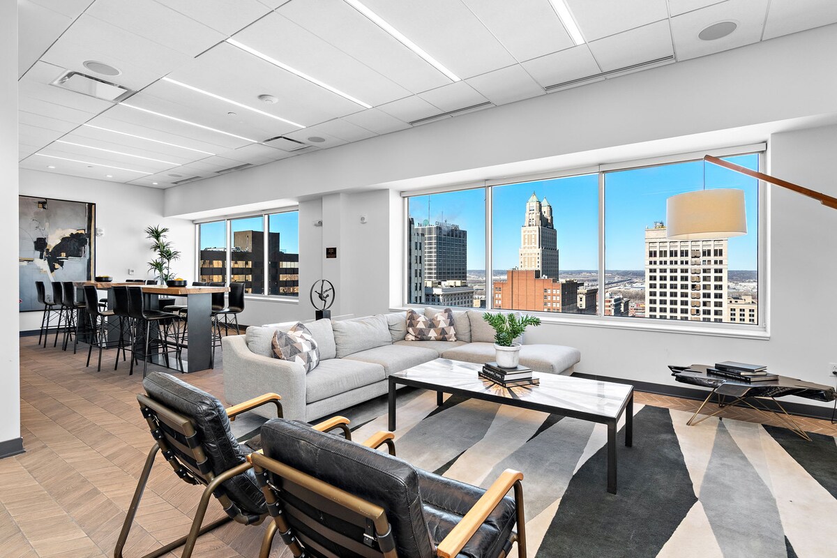 An open living area features large windows providing city views. The layout includes a comfortable sofa, coffee table, and black accent chairs. A dining table is visible in the background, surrounded by tall windows, enhancing the bright and inviting atmosphere of the space.