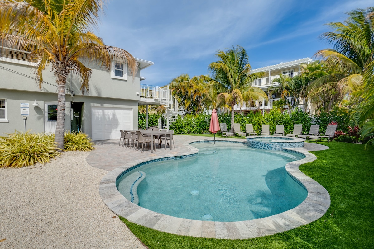 A spacious outdoor area features a heated pool with a sun shelf, surrounded by lounge chairs and a dining table. Lush greenery and palm trees provide shade, while a pink umbrella adds a pop of color. A sandy pathway leads toward the house, creating a welcoming environment.