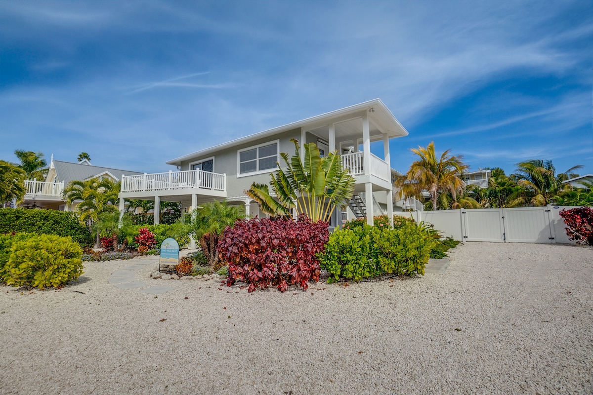 A two-story residence is situated on a landscaped property, surrounded by lush tropical plants and colorful shrubbery. The bright exterior is complemented by multiple balconies, creating inviting outdoor spaces. A gravel pathway leads to the entrance, enhancing the property's curb appeal in a sunny environment.