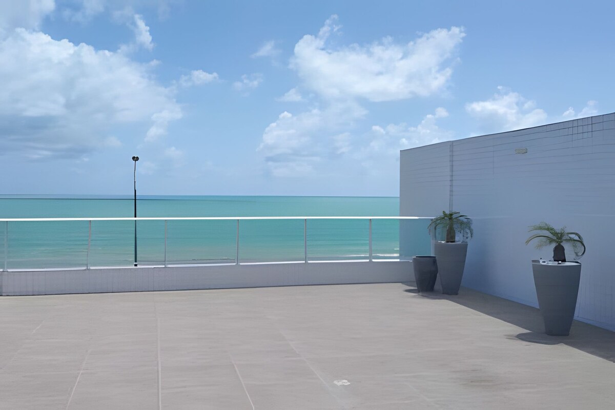 A spacious outdoor terrace showcases a smooth tiled floor with sea views in the background. Two large, modern planters are positioned at the edges, each containing a plant. The bright blue sky contrasts with the turquoise waters, enhancing the serene atmosphere.