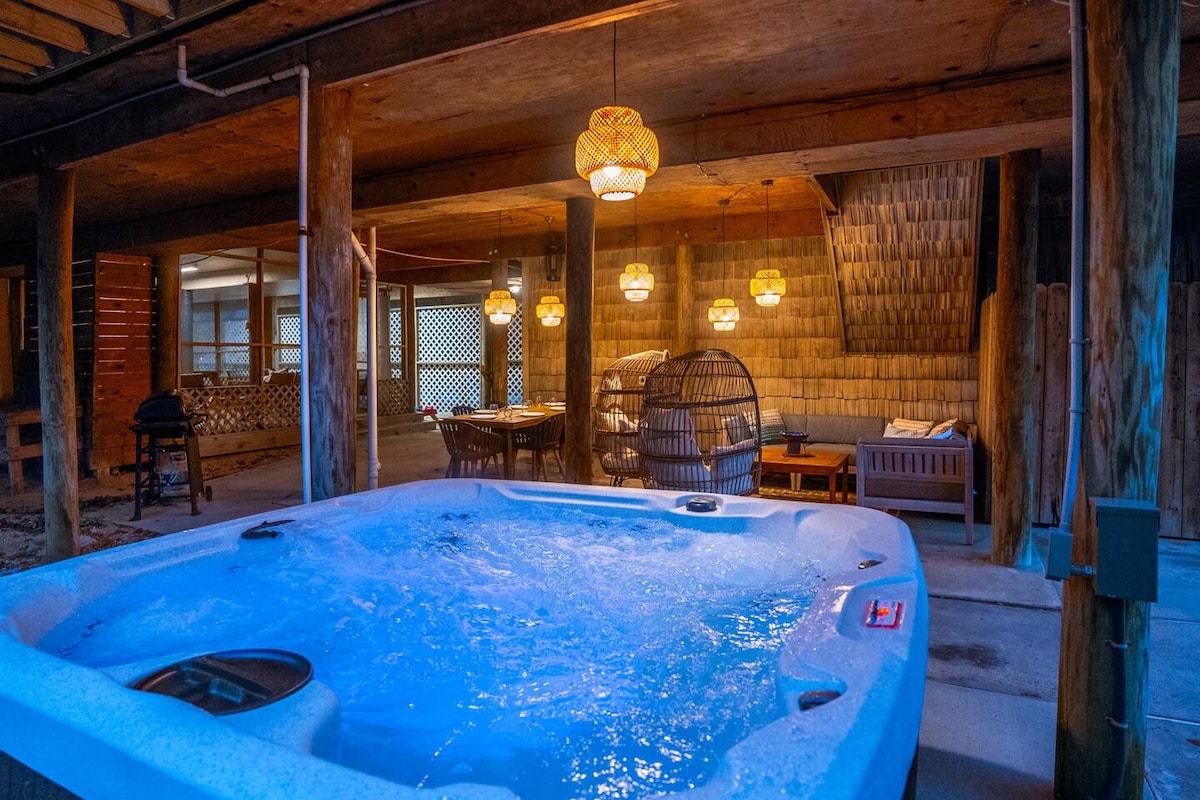 A spacious outdoor area features a hot tub surrounded by rustic wooden beams and soft ambient lighting from hanging fixtures. Cozy seating areas are visible in the background, providing a comfortable space for relaxation and gathering.