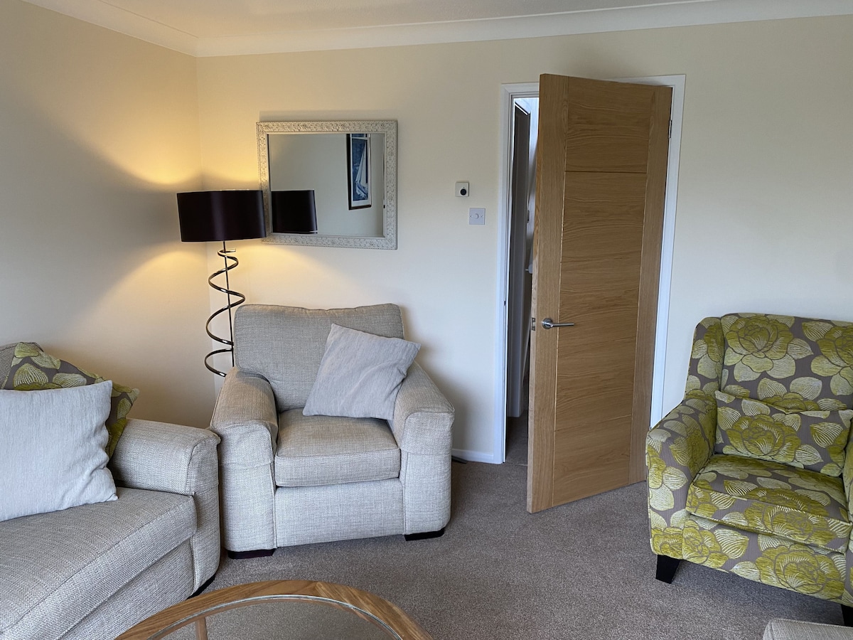 A comfortable lounge area features two armchairs and a sofa, with light-colored cushions enhancing the space. A decorative mirror reflects the room's ambiance, complemented by a standing lamp in the corner. A door leads to the adjacent area, providing access to the rest of the home.