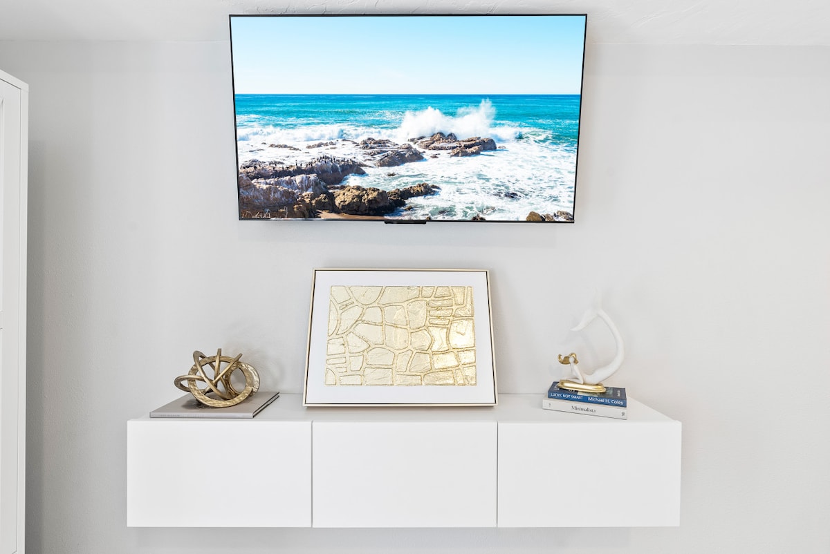A contemporary white media console is positioned below a wall-mounted television displaying a vibrant beach scene with crashing waves. An elegant framed artwork rests on the console alongside decorative objects, providing a touch of style to the living area.