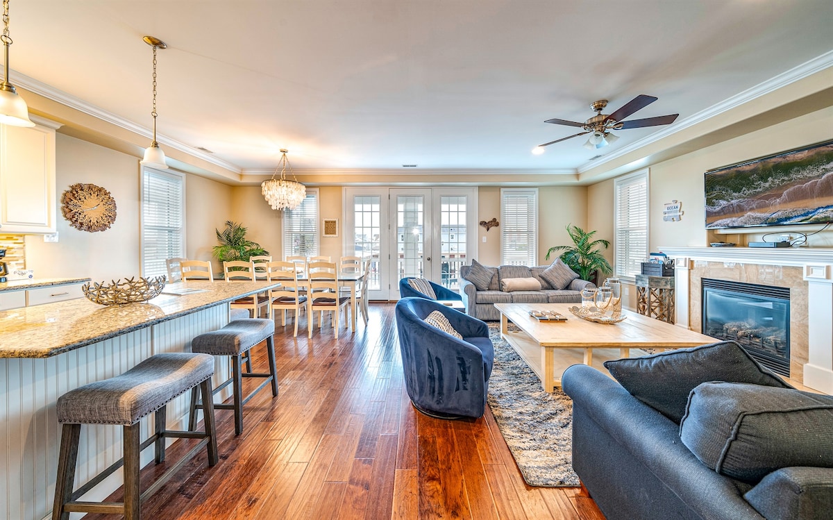 An open-concept living area features a cozy arrangement of plush sofas and accent chairs, centered around a wooden coffee table. A large entertainment unit with a flat-screen TV enhances the space. A dining area with a grand table and six chairs is visible, illuminated by natural light.