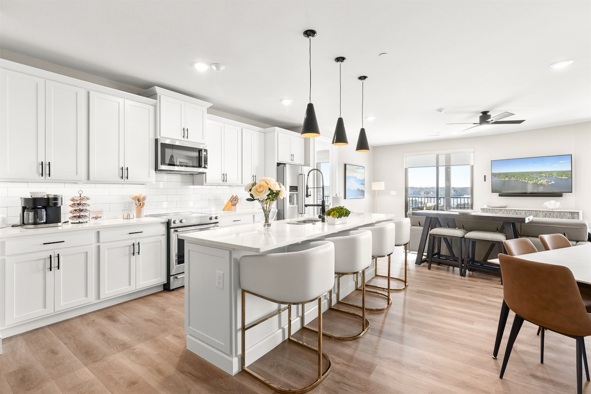 A modern kitchen area features ample white cabinetry and stainless steel appliances. A large central island with seating for six is complemented by three pendant lights above. Window views showcase the surrounding landscape, while a comfortable dining area is positioned nearby.