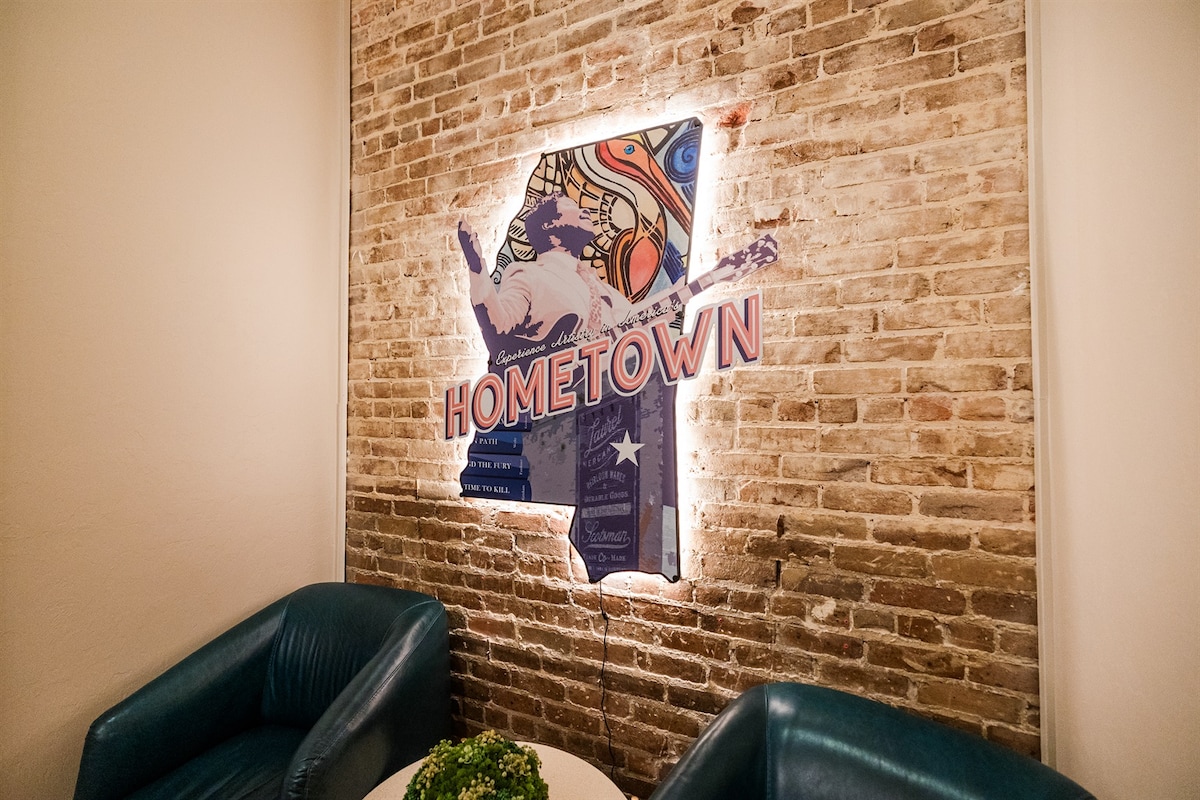 A decorative wall showcases an illuminated sign featuring the word "HOMETOWN" against a textured, exposed brick background. The sign is designed with vibrant colors and artistic elements, adding a local touch to the space. Two deep blue chairs are positioned in front of the wall.