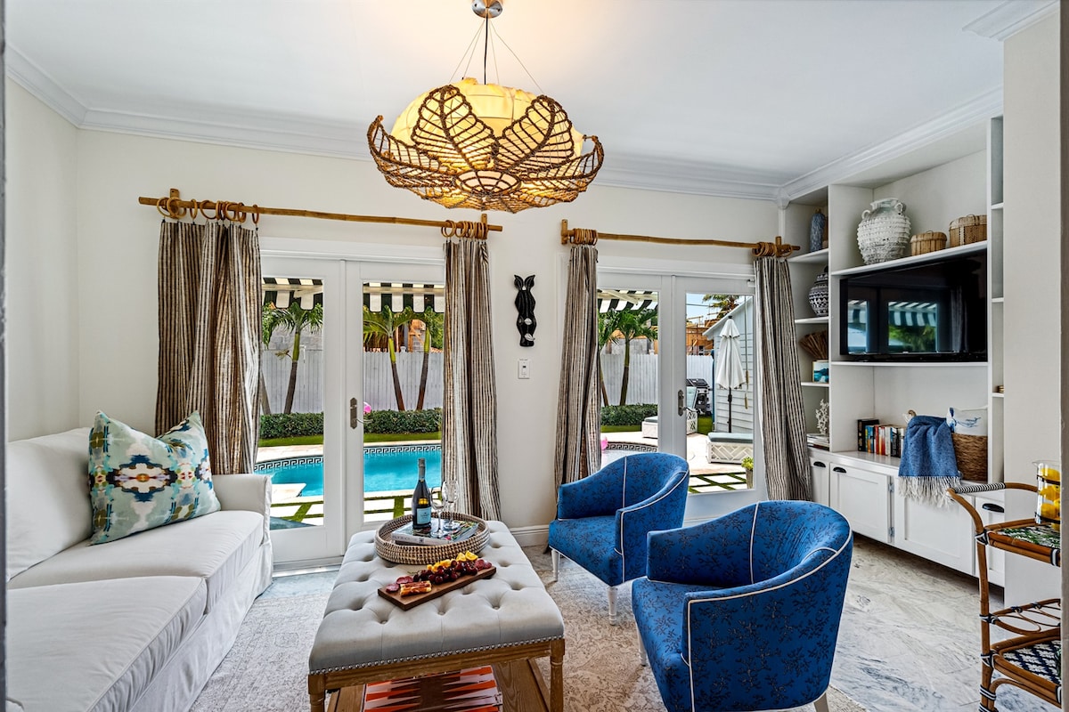 A bright sunroom features large glass doors that open to the outdoor pool area. A light-colored sofa with decorative cushions is complemented by two blue accent chairs. A textured coffee table displays a tray of snacks, while a stylish chandelier provides overhead lighting.