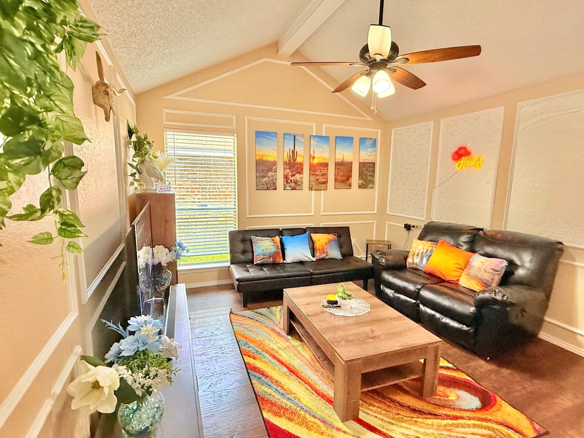 A bright living area showcases a black leather sofa and a pull-out sofa bed, with a wooden coffee table in the center. The vibrant rug adds warmth. Framed wall art displays scenic views, while natural light filters through a window with soft fabric treatments.