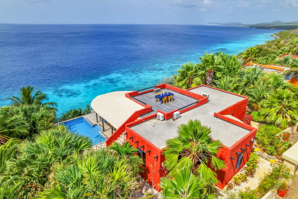 A stunning aerial view showcases Villa Kokopelli, highlighted by a vibrant red exterior. The private pool and sun terrace are visible, nestled among lush greenery, with the serene turquoise waters of the Caribbean Sea stretching out beyond.