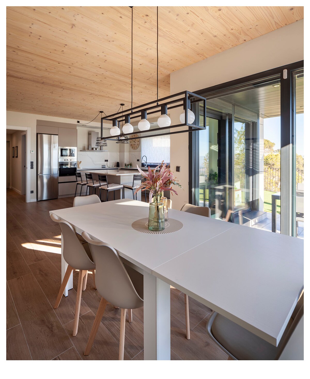 A modern dining area features a white table surrounded by light-colored chairs. Above, a minimalist light fixture holds five round bulbs. The open kitchen is visible in the background, alongside large glass doors leading to an outdoor terrace. Natural light fills the space.