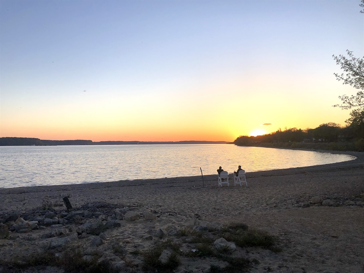 Two chairs are positioned on a sandy beach, facing a serene lake at sunset. The sky displays warm tones of orange and yellow as sunlight reflects on the water's surface. A tranquil shoreline and distant trees frame the background, creating a peaceful outdoor setting.
