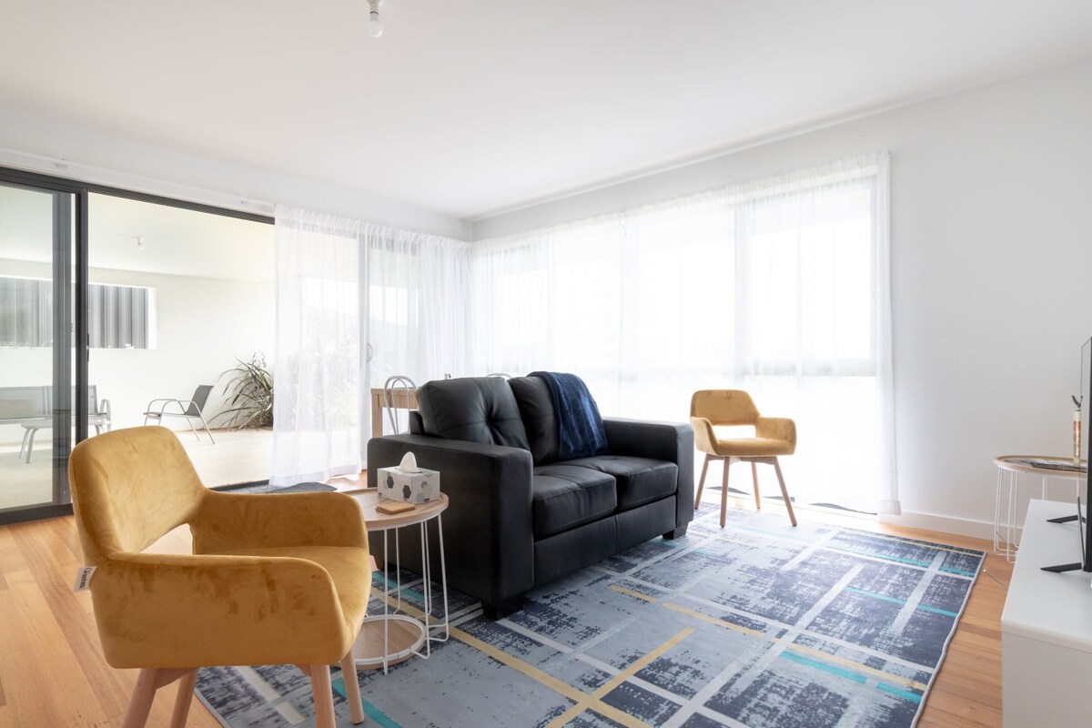 An open living space features a black sofa centered on a colorful geometric area rug. Two mustard-yellow armchairs are positioned nearby, and large glass doors provide an abundance of natural light. A simple coffee table sits beside the sofa, enhancing the cozy ambiance.