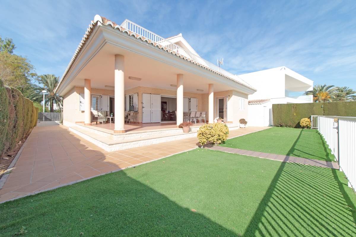 A spacious outdoor area features a well-maintained lawn surrounded by a hedge. A covered terrace provides seating for guests, with several chairs arranged around a table. Sunlight filters across the floor, highlighting the architectural details of the chalet.