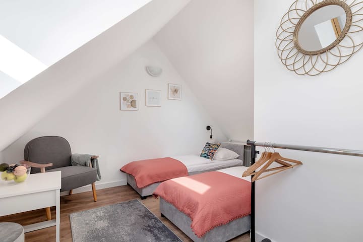 Druga spavaća soba s dva jednostruka kreveta, bijelom posteljinom s ružičastim detaljima i ukrasnim jastucima. Soba ima kos strop i na zidu je mali okrugli sat.