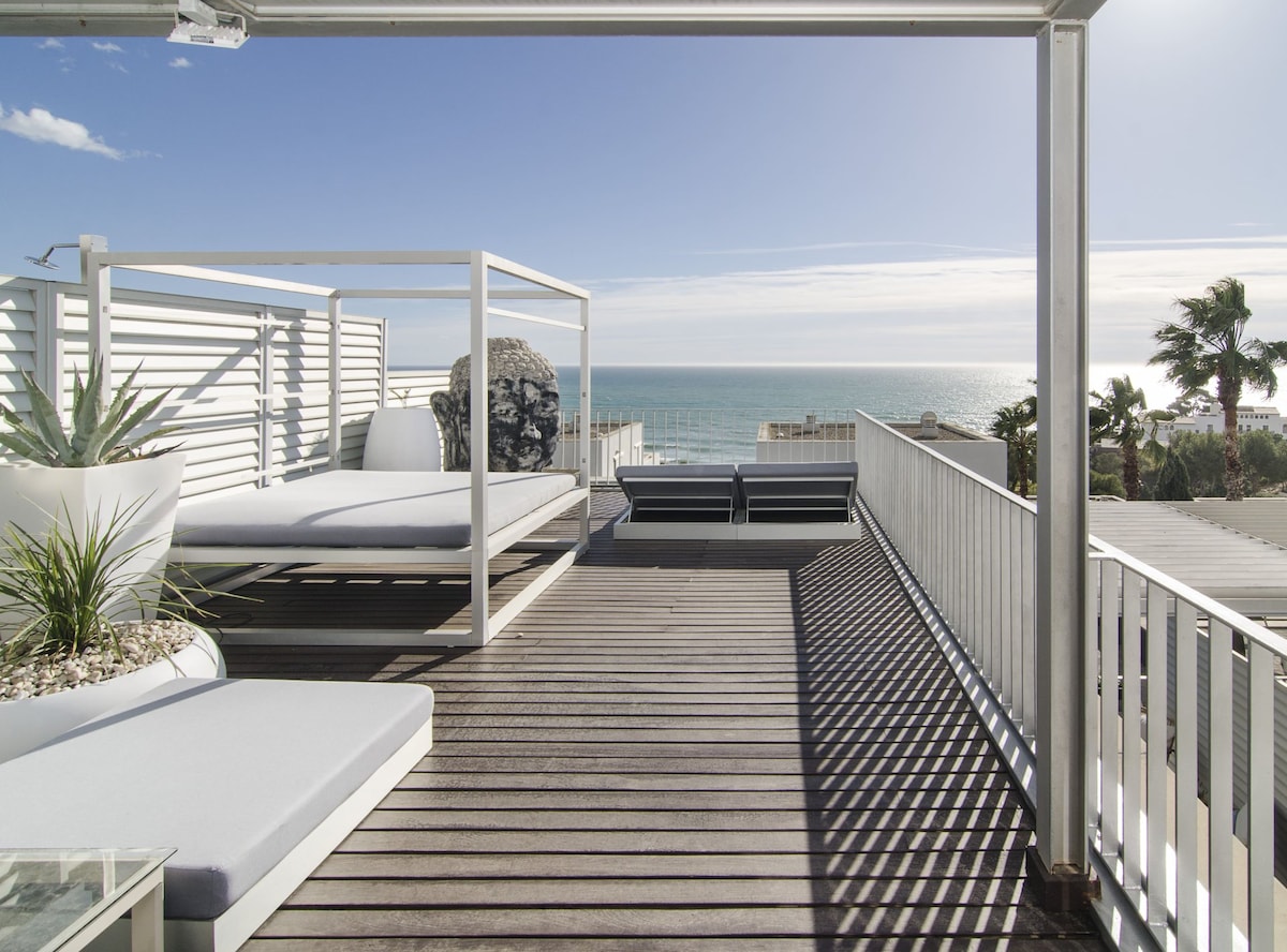 A spacious terrace is presented with comfortable lounge furniture and a view of the Mediterranean Sea. Decorative plants are positioned in planters, and bright sunlight creates a serene setting. The wooden decking offers an inviting surface for relaxation, with clear skies enhancing the expansive atmosphere.