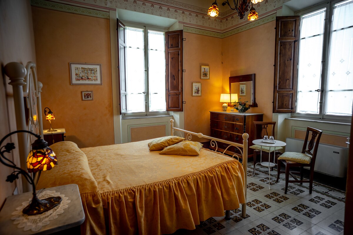 A delicate bedroom features warm, muted orange walls. A spacious bed with a soft yellow coverlet is complemented by two windows with wooden shutters. A wooden dresser and a small table with a chair create inviting spaces for relaxation and comfort.