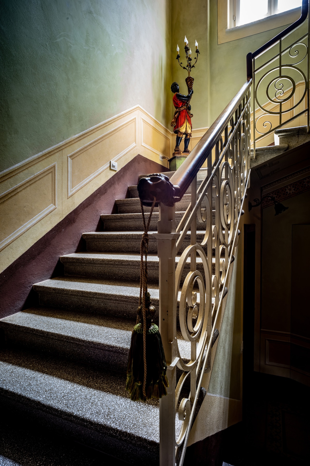 A staircase featuring a decorative railing ascends alongside a wall painted in soft green hues. Artifacts, including a tall statue, are positioned at the landing. Natural light filters through a window above, softly illuminating the space.