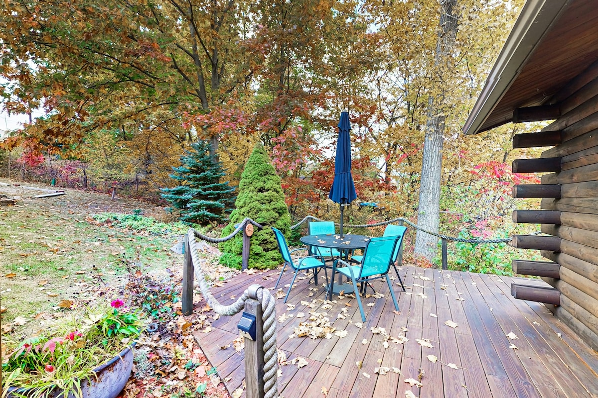 An inviting outdoor deck is furnished with a four-person dining table and chairs in vibrant blue. Surrounding the deck, trees display autumn foliage, and a tall umbrella offers shade. Decorative plants and a rope railing enhance the natural charm of the space.