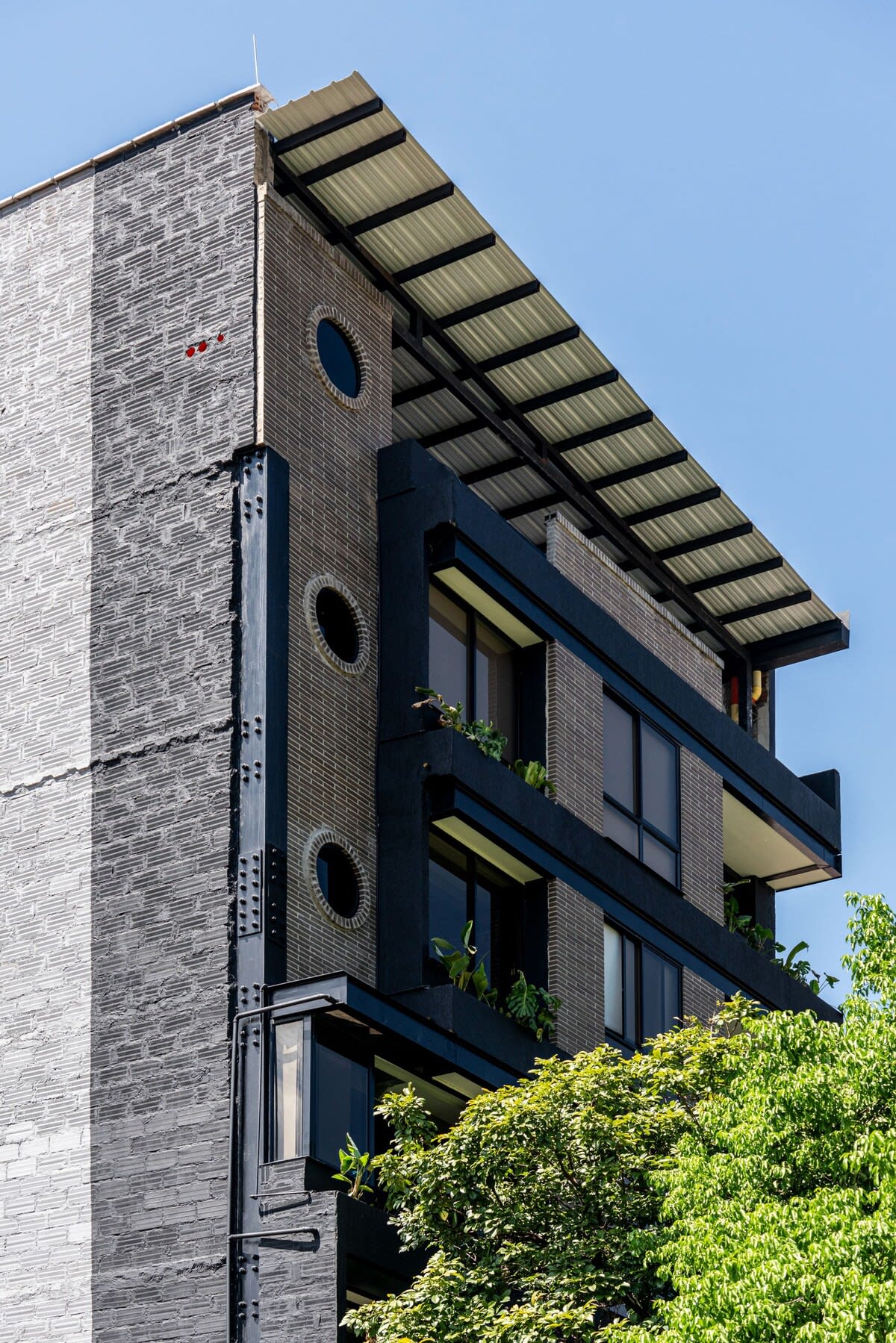 The exterior of a modern building is featured, showcasing a combination of textured gray walls and sleek black accents. Round windows enhance the architectural design. Lush greenery is visible, adding a touch of nature to the urban setting, under a clear blue sky.