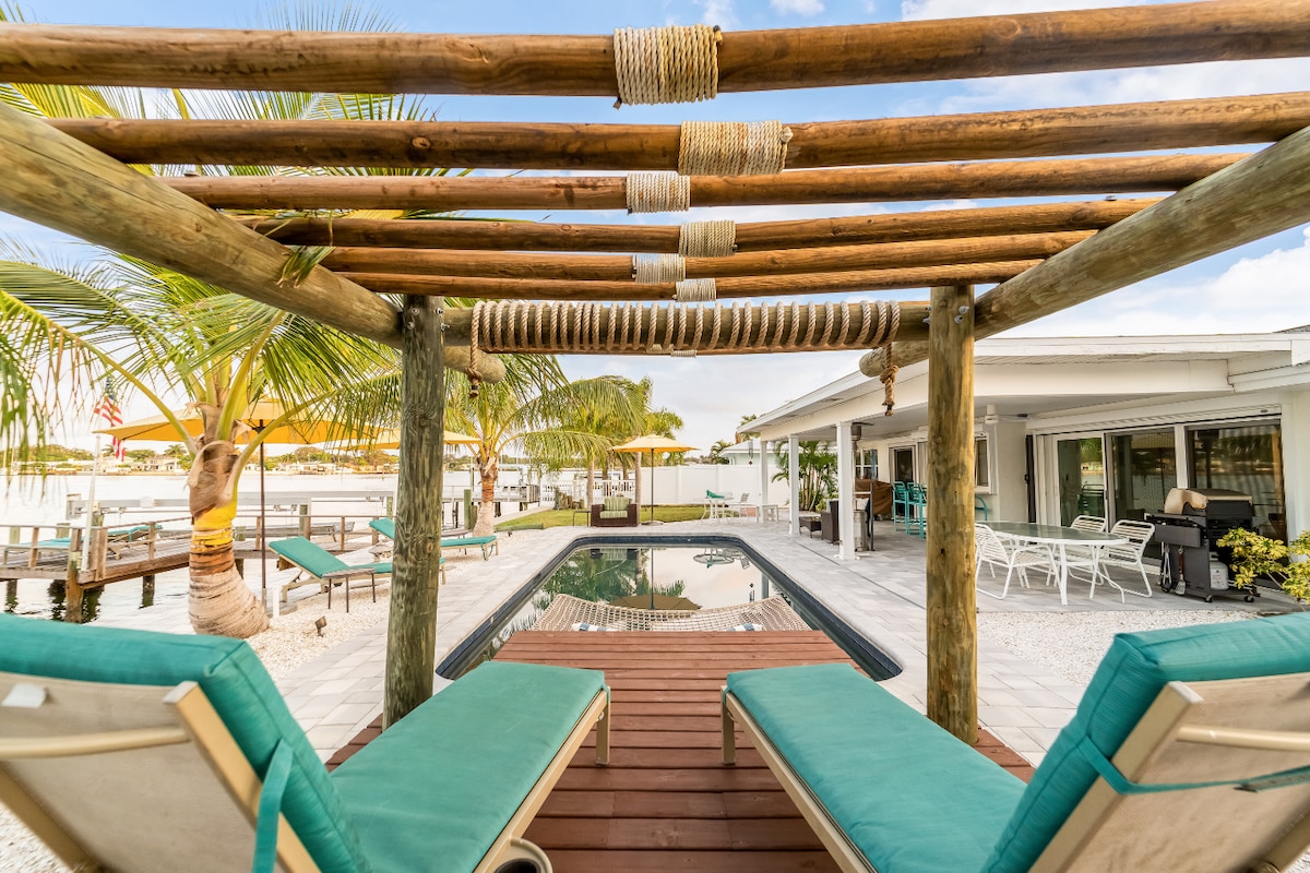 A tranquil pool area is framed by a wooden pergola with rope accents. Two lounge chairs with turquoise cushions are positioned beside the rectangular pool, reflecting the surrounding palm trees and outdoor seating areas. The serene waterfront view completes the inviting outdoor space.