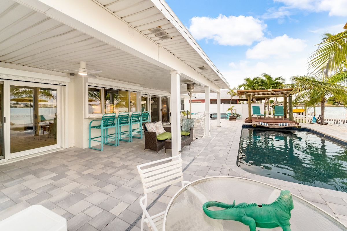 The outdoor patio area features a spacious layout with comfortable seating arrangements and high stools at a bar. A swimming pool is visible, surrounded by tropical landscaping and a gazebo. Large glass doors allow for unobstructed views of the waterfront beyond.