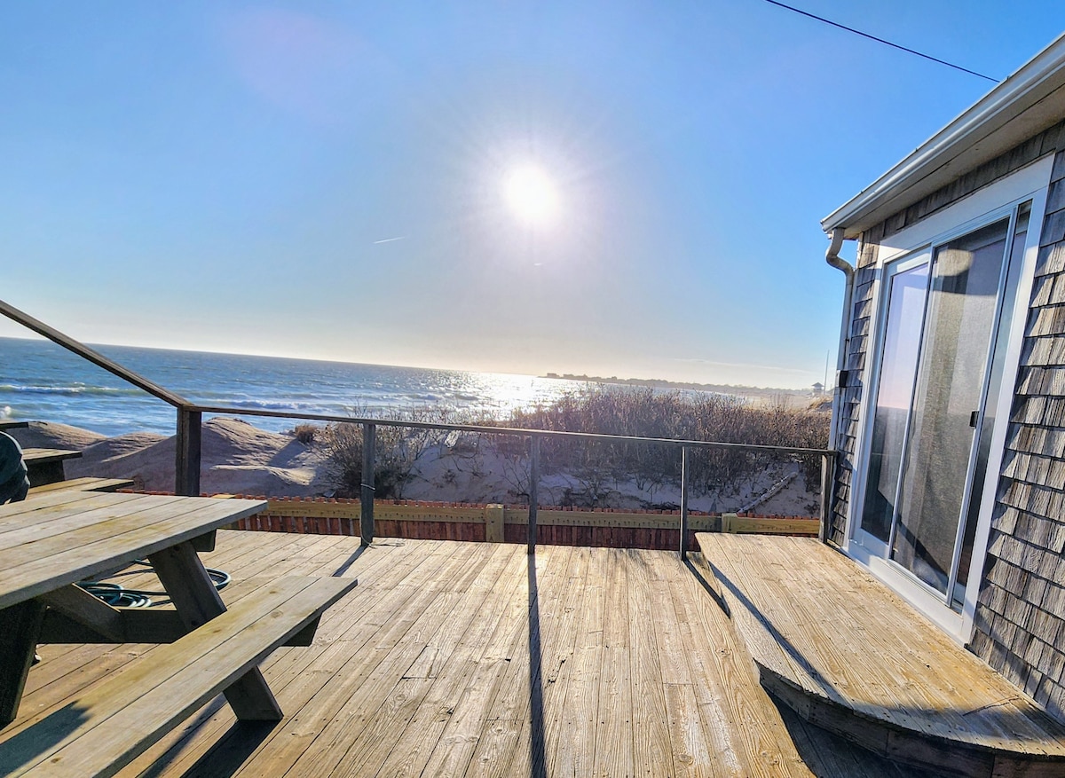 A sunlit wooden deck is visible, featuring a picnic table overlooking the ocean. Waves gently lap at the shore in the background under a bright sun. The horizon stretches in the distance, blending the blue sky with the water.