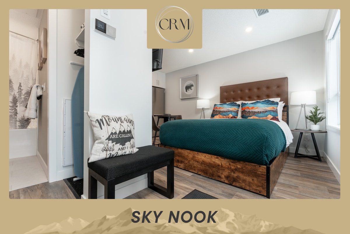 A spacious studio suite features a cozy Queen bed, adorned with soft bedding and decorative pillows. Natural light illuminates the modern decor, while a plush bench sits invitingly near the entrance. A well-appointed bathroom area is visible in the background, enhancing the thoughtful design of the space.
