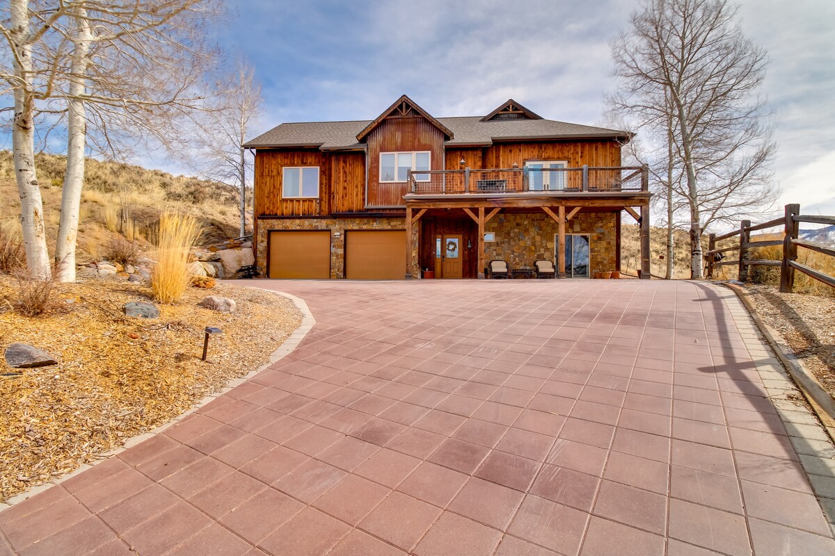 A spacious driveway leads to a two-story wooden structure, featuring large windows and a wraparound porch. Two garage doors are discernible, framed by decorative landscaping with rocks and low plants, while the surrounding hillside is dotted with bare trees.