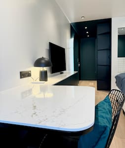 A contemporary workspace is set against a sleek wall, featuring a white marble table complemented by a black lamp. A television is mounted on the wall, while dark cabinetry provides storage. The room design emphasizes clean lines and a functional layout.