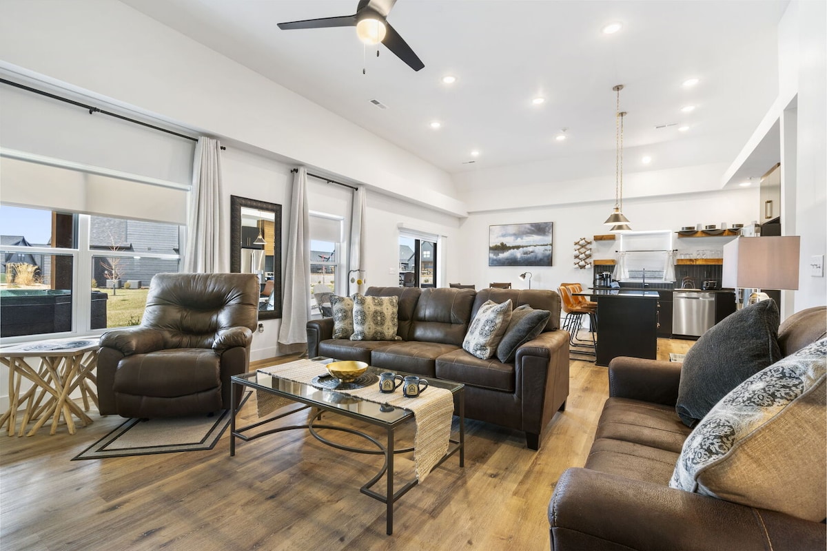 An open living room features comfortable seating arrangements, including leather recliners and a plush sofa. Large windows invite natural light, while a coffee table is centered in the space. The kitchen area is visible in the background, showcasing modern appliances and a dining area.