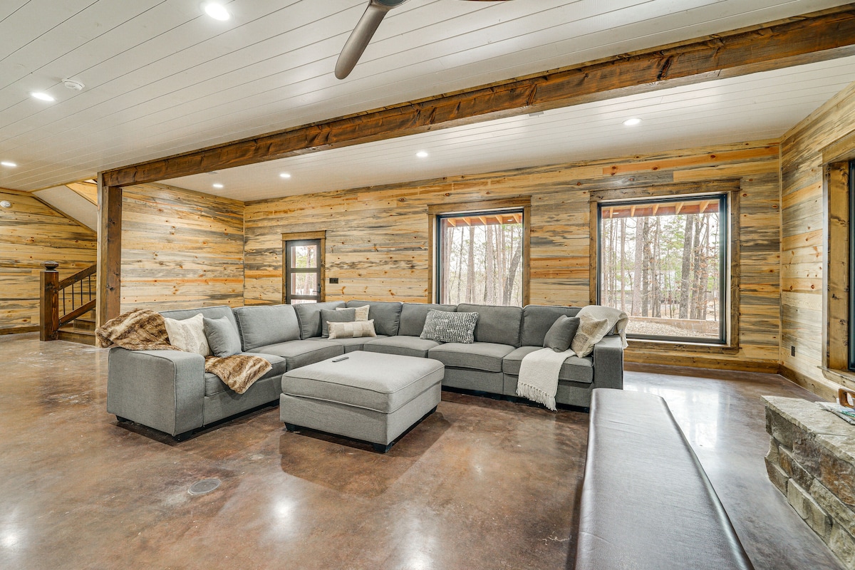 A spacious living area features a large sectional sofa with neutral tones, complemented by textured throws. Natural light filters through multiple windows, offering views of the surrounding trees. The rustic wooden walls and polished concrete floors provide a warm and inviting ambiance.