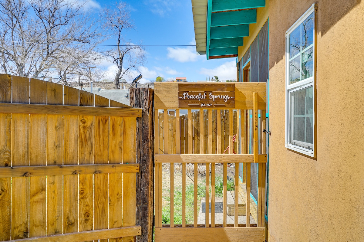A wooden gate adorned with the phrase 'Peaceful Springs' opens into a private yard. The gate is framed by a rustic wooden fence and leads to a well-maintained grassy area, gently illuminated by natural sunlight. A small window on the house is visible nearby.