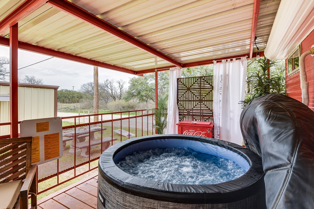 A covered porch features a private hot tub surrounded by scenic outdoor elements. The space is designed with wooden flooring and includes a backdrop of natural greenery, enhancing the feeling of seclusion.
