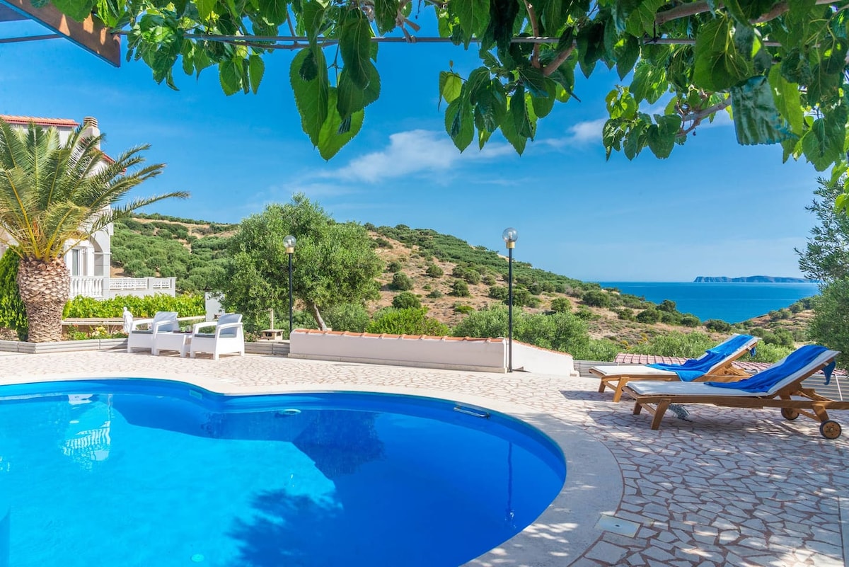 A clear blue pool is framed by a cobblestone patio, providing a view of the sea and rolling hills. Sun loungers and small seating areas are positioned nearby under leafy trellises, creating a serene outdoor setting.