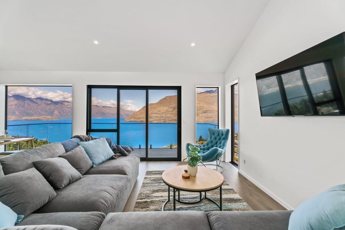 A modern living area is presented with a large sectional sofa adorned with decorative cushions. Floor-to-ceiling windows frame stunning views of Lake Wakatipu and surrounding mountains. A round coffee table rests on a textured area rug, while a comfortable chair adds to the inviting space.