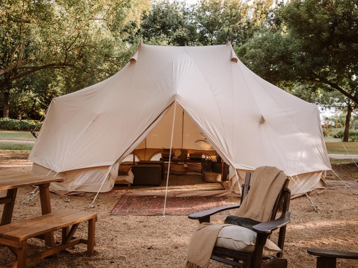 A spacious tent occupies a natural outdoor setting, featuring an open entrance that reveals a cozy interior. Nearby, a wooden table and chair are situated on a pebbled area, while trees provide ample shade and greenery surrounds the campsite.