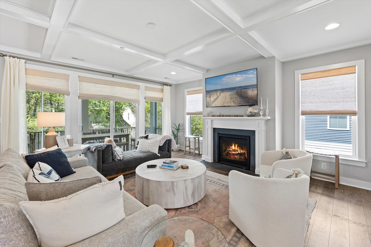 A spacious living area features a light-colored sofa and armchairs arranged around a circular coffee table. A modern fireplace is centered on the wall, with a large screen displaying coastal imagery above it. Natural light streams through large windows, illuminating the room.