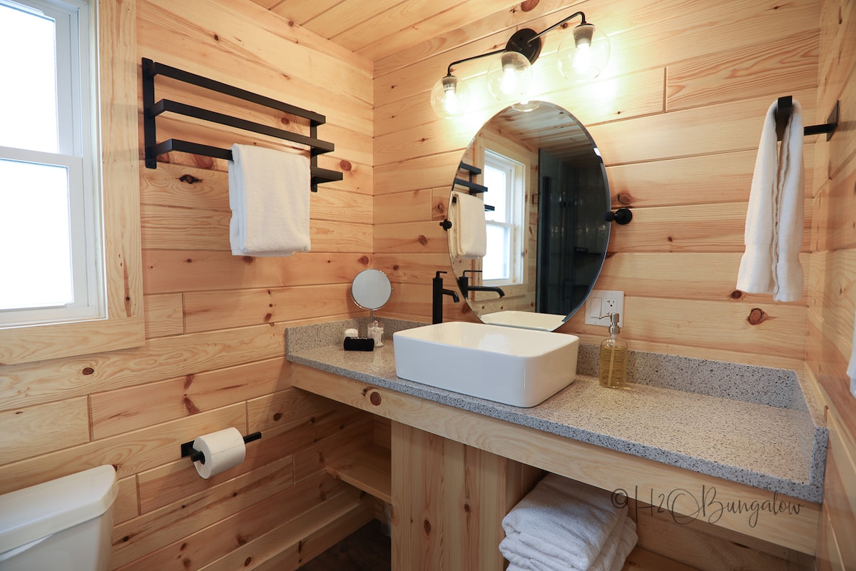A modern bathroom featuring natural wood paneling and a circular mirror above a sleek countertop. A clean white sink sits atop the surface, with warm LED lighting accentuating the design. Soft towels are neatly hung, enhancing the organized and functional space.