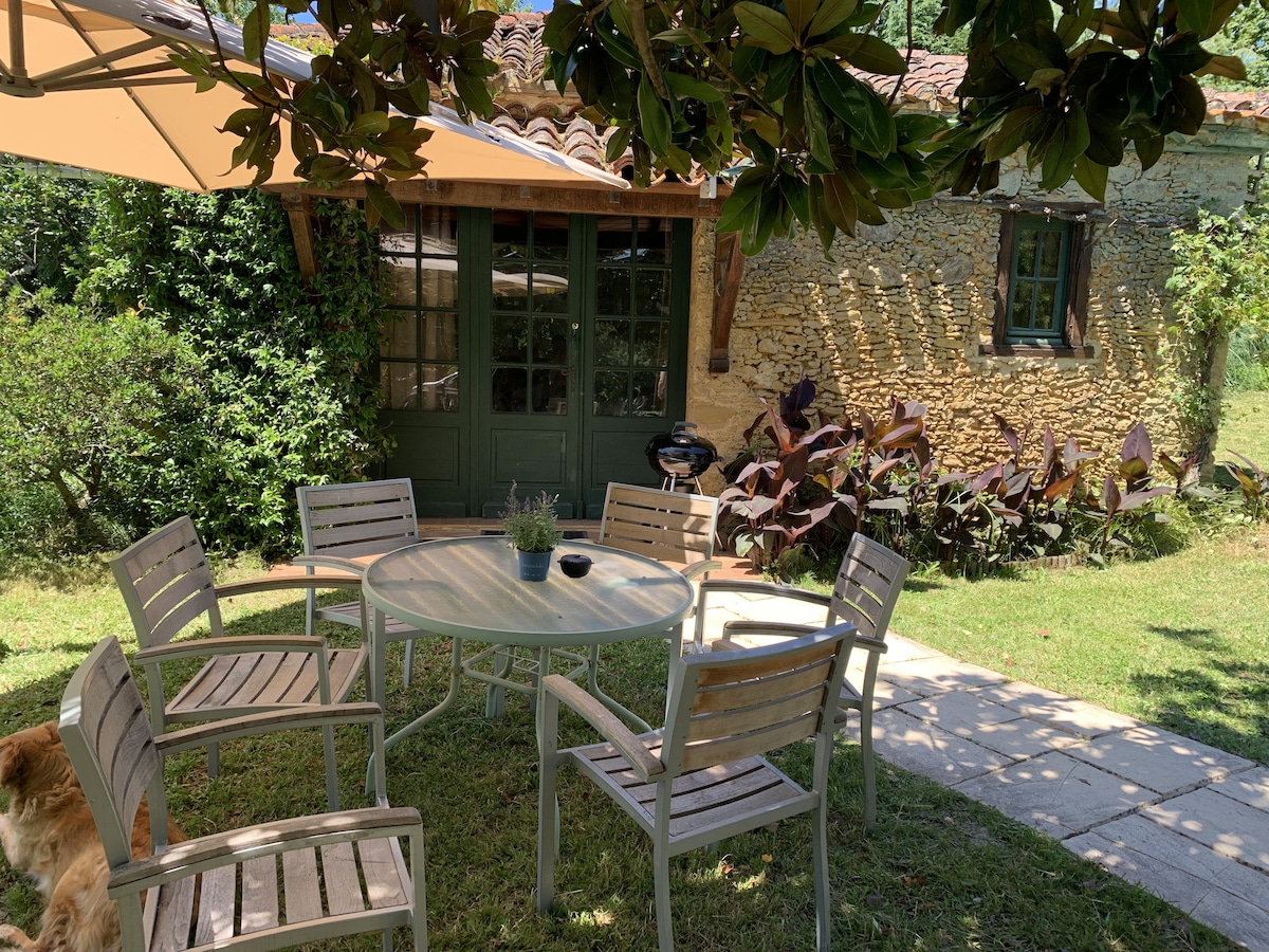 An outdoor seating area is featured, surrounded by lush greenery. A round table with several wooden chairs is positioned under a sunshade, providing a comfortable spot for relaxation. The rustic stone wall of the cottage is visible in the background.