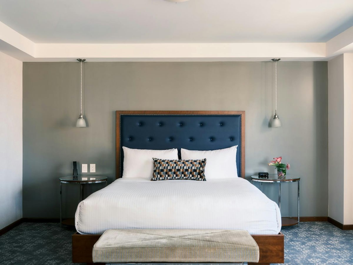 A spacious bedroom is highlighted by a plush queen bed with a tufted navy headboard. Flanking the bed are two round side tables with stylish lamps. A neutral-colored bench sits at the foot of the bed, complemented by soft carpeting and minimalistic decor.
