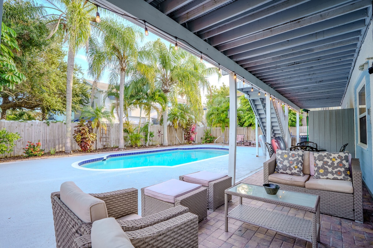 An outdoor patio area features a seating arrangement with plush cushions and a glass coffee table, set against a backdrop of palm trees. A shared heated pool is visible, surrounded by a mix of tropical foliage, creating a relaxed and inviting environment.