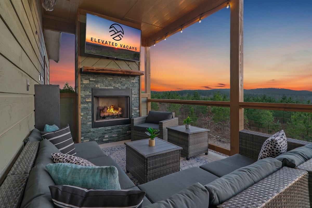 An inviting outdoor seating area is displayed on the balcony, featuring plush sectional sofas surrounding a stone fireplace. A mounted TV is visible above the fireplace, while string lights create an ambiance. The backdrop showcases a vibrant sunset over the forested mountains.